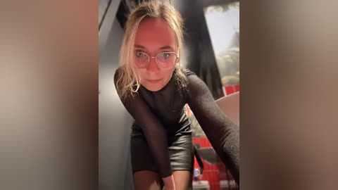 Media: Video of a blonde woman with glasses and a black leather dress, leaning forward with a confident expression, taken from a low angle, indoors with blurred background.