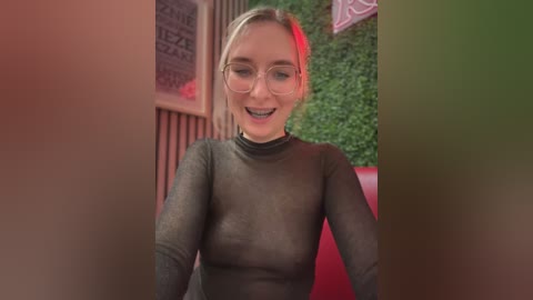 Media: A video of a smiling, fair-skinned woman with blonde hair and glasses, wearing a sheer black top, standing indoors with greenery and red accents in the background.