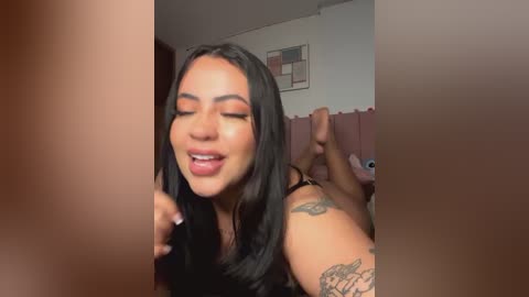 Media: Video of a smiling Latina woman with long black hair, wearing makeup, and a black top, with a tattoo on her arm, in a bedroom with pink curtains.