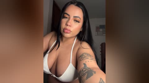 Media: A video of a Latina woman with long black hair, wearing a white bikini top, standing in a dimly lit room. She has a tattoo on her left arm.