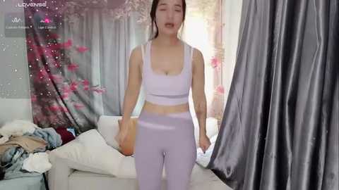 Media: A video of an Asian woman with fair skin and medium-length brown hair, wearing a light purple sports bra and matching leggings, standing in a bright, sunlit room with a floral tapestry and a white couch.