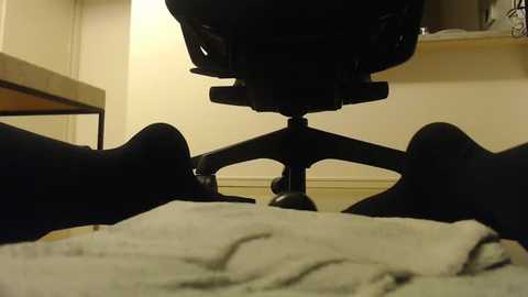 Media: Video of a person's legs wearing black socks, resting on a gray blanket on a beige floor. A black office chair with a swivel base is visible in the background against a cream-colored wall.