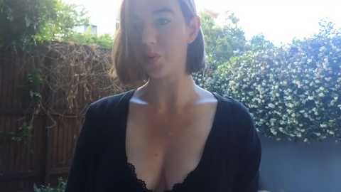 Media: Video of a fair-skinned woman with short, light brown hair wearing a black lace top, standing in a garden with a wooden fence and greenery in the background.