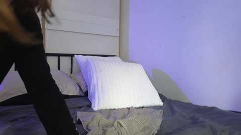 Media: Video of a woman in black pants, standing on a bed with white pillows and gray bedding, in a dimly lit room with purple light.