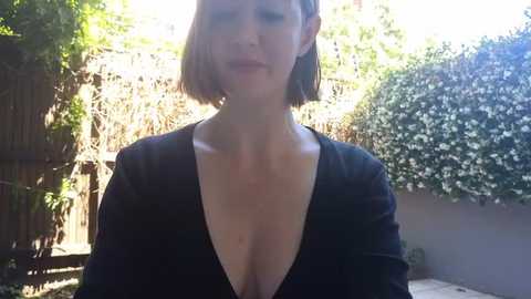 Media: Video of a light-skinned woman with short, dark brown hair, wearing a black V-neck top, standing in a sunlit garden with lush greenery and a wooden fence.