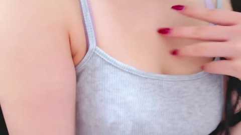 Media: Video of a woman with light skin, wearing a light gray, ribbed tank top that reveals her ample cleavage. She has a hand with bright red nail polish on her left breast, suggesting a playful or intimate gesture.