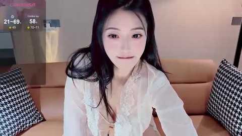 Media: A video of an East Asian woman with long black hair, wearing a sheer white lace top, sitting on a tan leather couch with black and white houndstooth pillows in a modern, dimly-lit room.