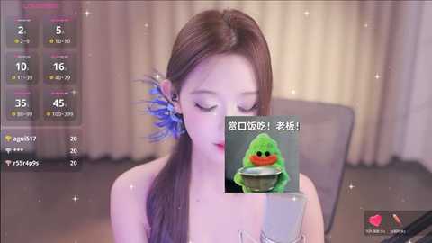 Media: A video of an Asian woman with long brown hair and blue hair accessories, wearing a green frog costume, sitting at a table.