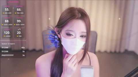 Media: Video of an East Asian woman in a white mask, with long brown hair, wearing a black top, sitting indoors with a blurred background, showing social media stats.