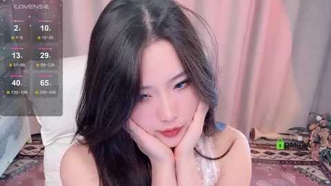 Media: Video of an East Asian woman with long black hair, fair skin, and full lips, wearing a white lace bra, resting her head on her hands. Background includes a plush chair and a cluttered desk.