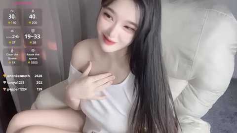 Media: A video of an East Asian woman with long black hair and fair skin, wearing a white off-shoulder top, sitting on a white cushion, smiling.