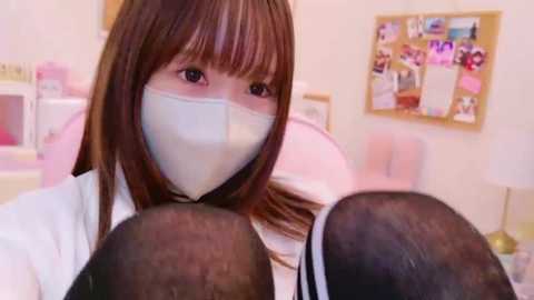 Media: Video of a young Asian woman with long brown hair, wearing a white face mask and a white lab coat, sitting in a brightly lit room with a bulletin board and pink furniture.