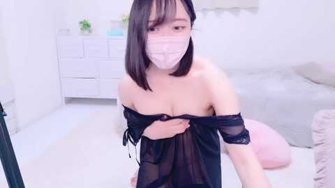 Media: Video of an East Asian woman with straight black hair, wearing a black sheer nightgown, a pink surgical mask, and a face shield. She is kneeling in a minimalist, bright bedroom with a white dresser and a gray bed.