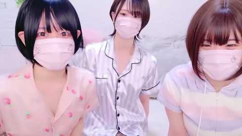 Media: Video of three young Asian women wearing white surgical masks, sitting on a bed. They have short black hair, and are dressed in white satin pajamas with black trim, revealing their bare chests. The background is a white room with minimal decor.