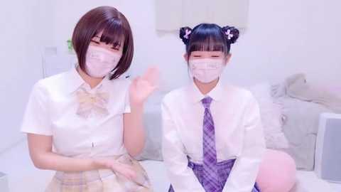 Media: Video of two Japanese schoolgirls in white blouses, light pink bow ties, and face masks, sitting on a bed. They have bobbed hair with one wearing twin buns and the other straight.