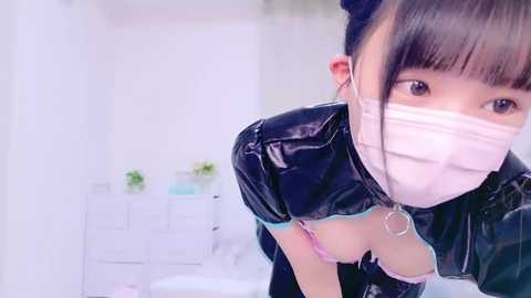 Media: A video of an Asian woman with straight black hair and a surgical mask, wearing a shiny black latex dress with a revealing neckline, leaning forward in a sterile, white-walled room with green plants and medical equipment.