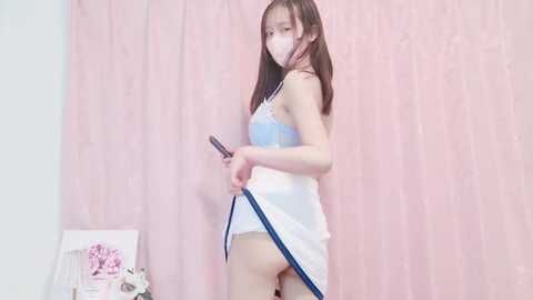 Media: A video of an Asian woman with long brown hair and light skin, wearing a white camisole and blue panties, holding a blue belt. The background features pastel pink curtains and a white chair with a pink bow.