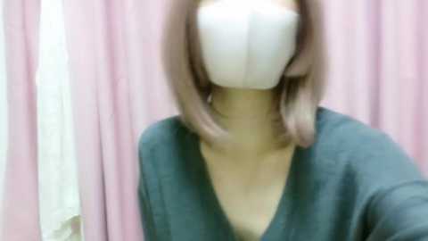 Media: A blurry video of a person with shoulder-length, light brown hair wearing a white face mask and a dark green V-neck sweater. The background features pink curtains and a white wall.