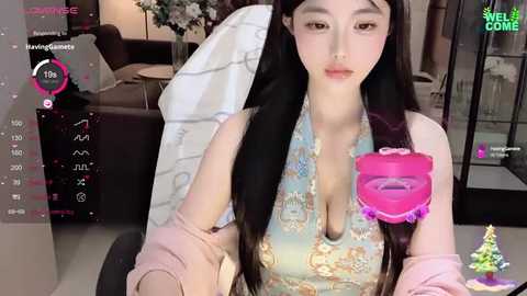 Media: Video of an Asian woman with long black hair, wearing a floral halter top, holding a pink heart-shaped box, seated in a modern living room.