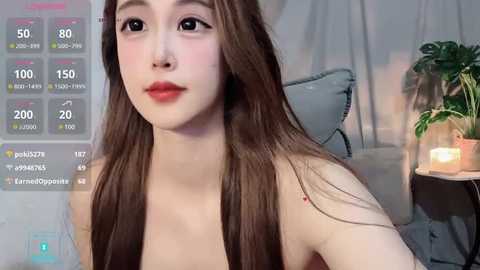 Media: Video of an East Asian woman with long brown hair, fair skin, and full red lips, wearing a strapless top, sitting in a cozy room with a lit candle, gray cushion, and potted plant.