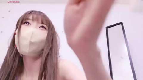 Media: Video of an Asian woman with long brown hair, wearing a white face mask and a black top, looking up at a mirror, in a minimalist white room with a black-framed mirror.