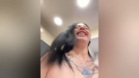 Media: A candid video of a smiling, topless woman with long black hair, wearing a blue necklace, in an indoor setting with beige walls and ceiling lights.