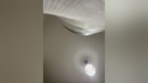 Media: Video of a ceiling light with a textured white shade, emitting a soft, diffused light, positioned in a dimly lit room with beige walls.