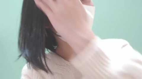 Media: Video of a woman with light skin and straight black hair, wearing a white sweater, with her hand covering her mouth, against a plain green background.