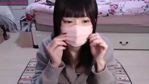 Media: Video of an Asian woman in a pink face mask, kneeling on a striped rug in a pastel bedroom, adjusting the mask with both hands.