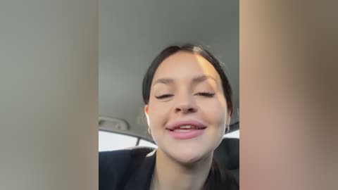 Media: Video of a woman with light skin, dark hair, and closed eyes, smiling widely, taken from a low angle, possibly in a car, with blurred background.