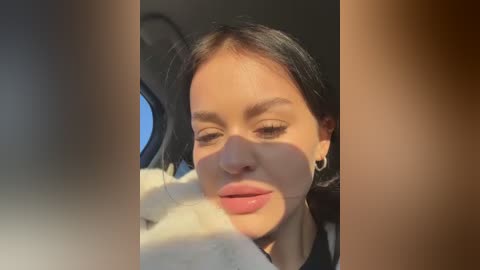 Media: Video of a woman with long dark hair, fair skin, and light pink lipstick, wearing a white top, sitting in a car with sunlight streaming in, creating a warm, natural glow.