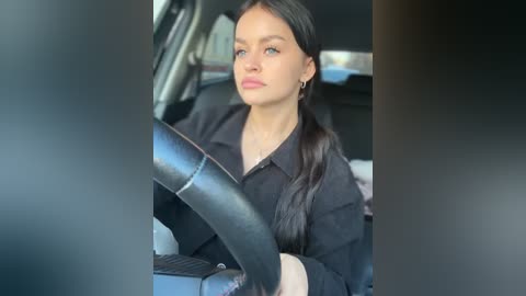 Media: Video of a young woman with long black hair, wearing a black shirt, driving a car, with a blurred background.