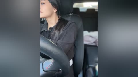 Media: Video of a young woman with fair skin, dark hair, and light makeup, wearing a black jacket, driving a car with a black interior. The background shows a messy, cluttered passenger seat.