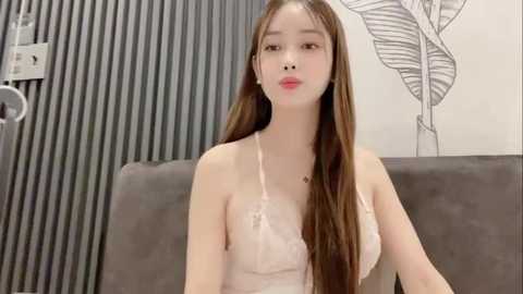 Media: Video of an East Asian woman with long, straight brown hair, wearing a light pink lace camisole. She sits on a gray sofa in a modern room with vertical gray-striped wallpaper and a black-and-white leaf pattern on the wall.