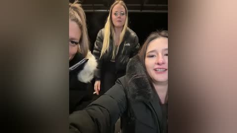 Media: Video of three blonde women in black coats, one smiling, one serious, and one partially obscured, in a dark room.