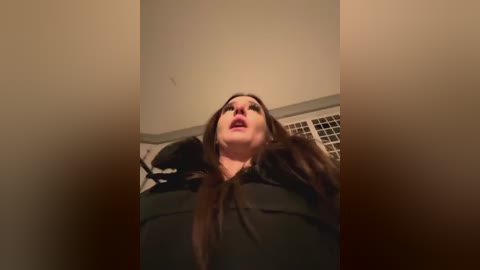 Media: A video captures a woman with long brown hair and fair skin, wearing a black blouse, looking upwards with an expression of surprise or shock, in a dimly lit room with beige walls and a tiled window.