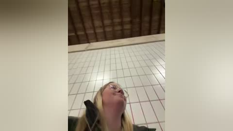 Media: Video of a woman with long blonde hair, wearing glasses, standing on a tiled floor, looking up at a brick ceiling.