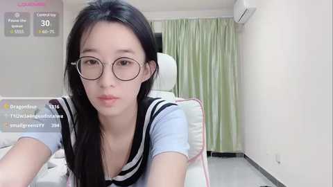 Media: Video of an East Asian woman with fair skin, long black hair, and round glasses, wearing a sailor-style school uniform, sitting on a white chair in a minimalist room with light green curtains and air conditioning.