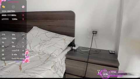 Media: Video of a modern bedroom with a dark wood headboard, white bedding with floral patterns, and a nightstand with a phone and electrical outlets.