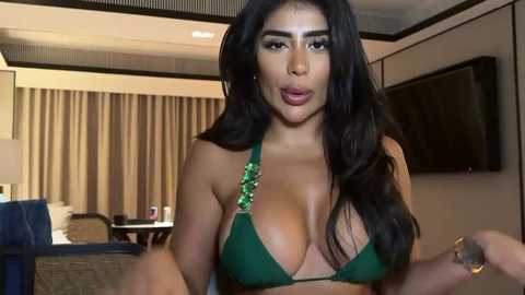 Media: Video of a voluptuous Latina woman with long black hair, wearing a green bikini top with a large jewel, posing indoors near a beige couch and curtains.