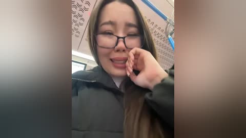 Media: A video of a young woman with fair skin, long brown hair, and glasses, smiling and playfully touching her mouth, wearing a black jacket. She's indoors, possibly on a bus, with a patterned ceiling in the background.