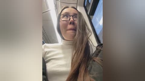 Media: Video of a Caucasian woman with long, light brown hair, wearing black-rimmed glasses and a white turtleneck, seated on a subway car. The background features subway car interior with seats and windows.