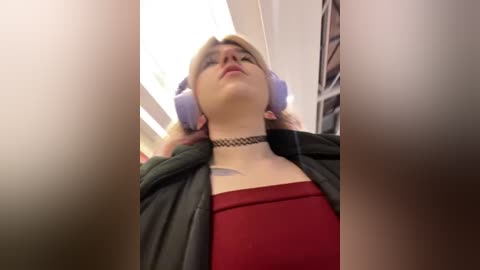 Media: A video of a woman in a red top, wearing a black choker and large purple headphones, with a blurred background, taken from a low angle.