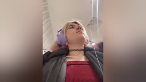 Media: A video of a young woman with platinum blonde hair, wearing a black choker necklace, gray hoodie, and red top, lying back in a bathtub with lavender headphones on, listening to music.