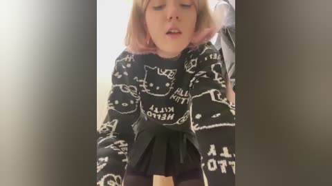 Media: A video of a young, light-skinned woman with shoulder-length blonde hair, wearing a black sweater with white cat illustrations, leaning forward suggestively.