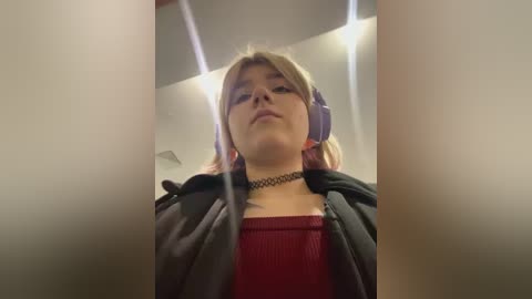 Media: Video of a young woman with light skin, wearing a red top and black choker, headphones, and a black hoodie, in an indoor setting with ceiling lights.