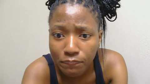 Media: Video of a young Black woman with medium-brown skin and braided hair, wearing a black tank top, looking worried against a plain white background.