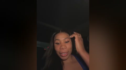 Media: Video of a Black woman with medium brown skin, wearing a dark top, adjusting her hair with her left hand. The background is dimly lit, creating a moody, intimate atmosphere.