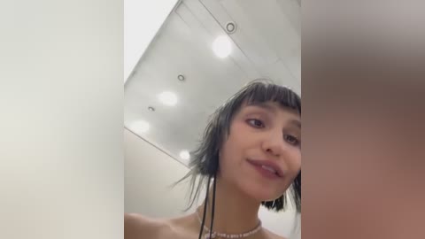 Media: Video of a young woman with short, dark hair and blunt bangs, wearing a black top, standing in a modern, white room with recessed ceiling lights.