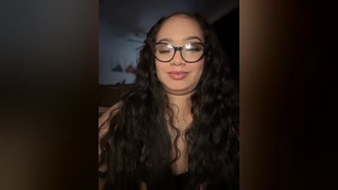 Media: Video of a smiling, light-skinned woman with long, curly black hair and glasses, wearing a black top, in a dimly lit room with blurred background.